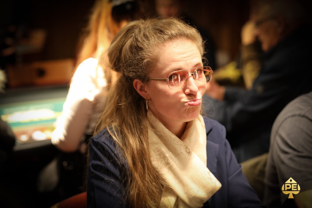 Ein Mann in einem schwarzen Anzug und einer roten Krawatte sitzt an einem Pokertisch mit grünen Filz. Er hält Karten in der Hand und schaut konzentriert auf den Tisch. Im