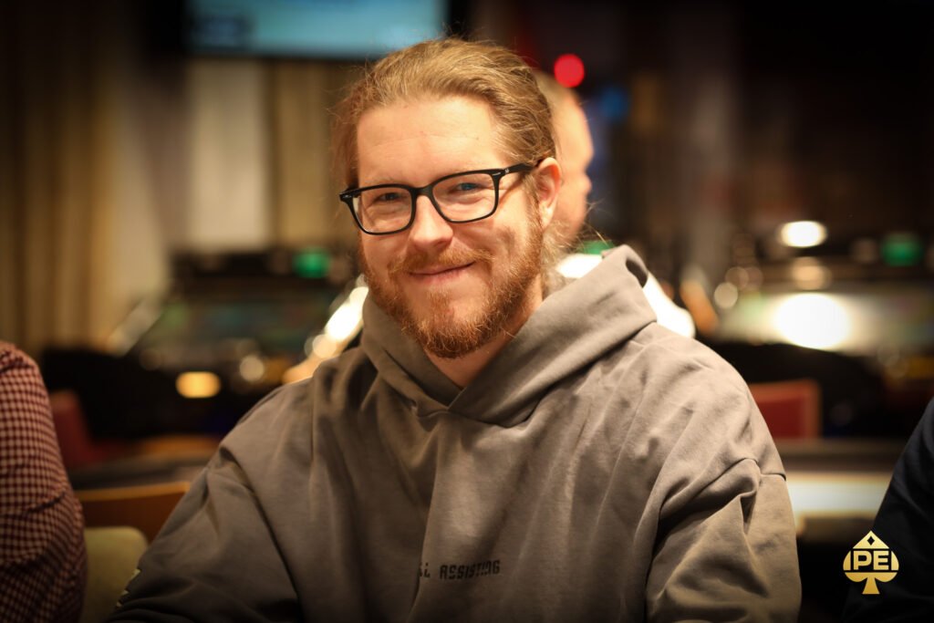 Ein Mann und eine Frau sitzen an einem Pokertisch und spielen eine Partie Poker. Der Mann hält Karten in der Hand und schaut konzentriert auf den Tisch, während die Frau ebenfalls Karten in der Hand h