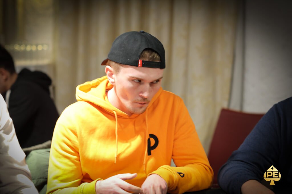 Ein Mann in einem dunklen Anzug, der eine Hand mit Pokerkarten hält, sitzt an einem Pokertisch in einem eleganten Casino. Im Hintergrund sind verschwommene Lichter und andere Spieler zu sehen.
