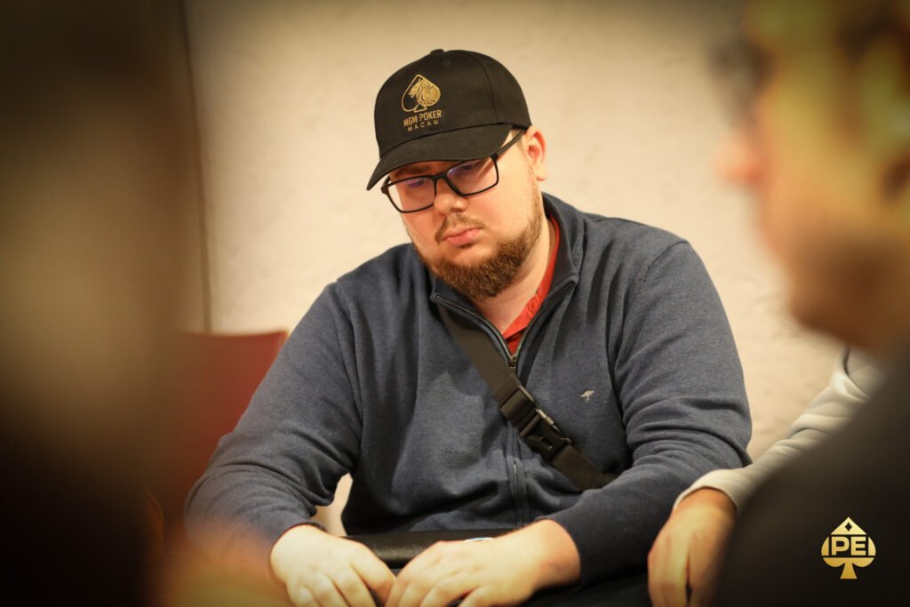 Ein Mann in einem dunkelblauen Anzug und einer weißen Krawatte sitzt an einem Pokertisch und hält seine Karten in der Hand. Im Hintergrund sind weitere Spieler und Pokerchips zu sehen.