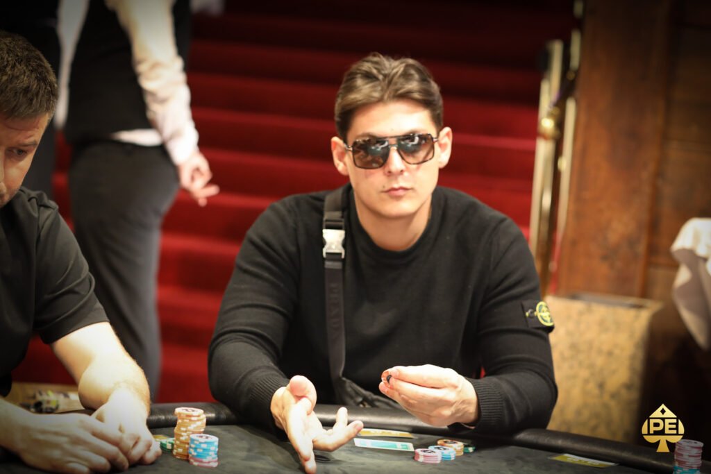 Ein professioneller Pokerspieler sitzt an einem Tisch mit Chips und Karten vor sich. Er konzentriert sich auf sein Blatt und betrachtet seine Gegner mit einem ernsten Gesichtsausdruck.