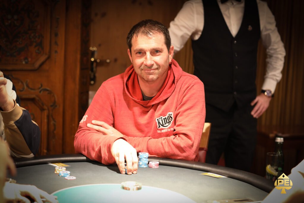 Ein Mann in einem dunkelblauen Anzug und einer Fliege steht vor einem Pokertisch mit Spielkarten und Chips. Er hält zwei Spielkarten in der Hand und schaut konzentriert auf den Tisch.