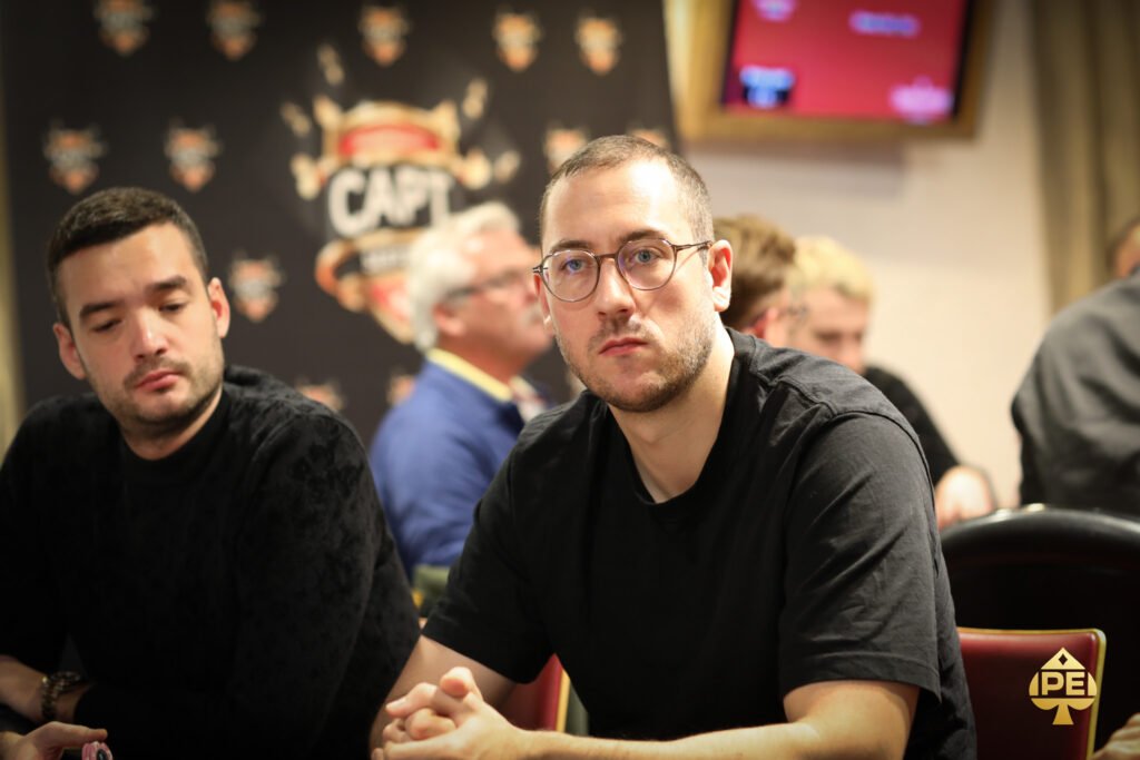 Ein Mann und eine Frau sitzen am Pokertisch und scheinen konzentriert auf ihre Karten zu schauen. Die Frau hält eine Hand mit Spielchips, während der Mann eine Zigarre raucht. Im Hinter