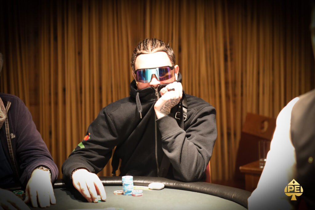 Ein Mann in einem dunkelblauen Anzug und einer weißen Hemd, der lächelnd vor einem pokertisch sitzt und Karten hält, während er auf seine Gegner schaut. Die Karten und Chips auf