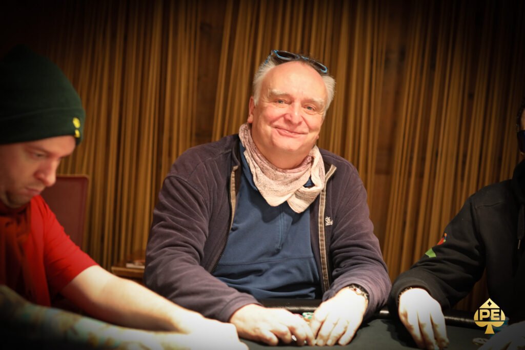 Ein Mann in einem schwarzen Anzug und einer schwarzen Sonnenbrille spielt Poker an einem Tisch mit grünem Filz. Er hält Karten in der Hand und betrachtet sie konzentriert, während andere