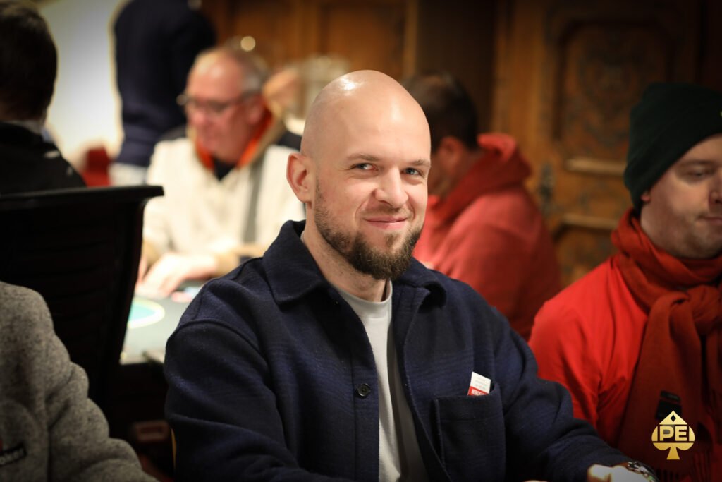 Ein Mann in einem eleganten Anzug sitzt an einem Pokertisch und hält seine Karten. Im Hintergrund sind weitere Spieler und eine Dealerin zu sehen. Die Spannung des Pokerspiels ist deutlich spürbar