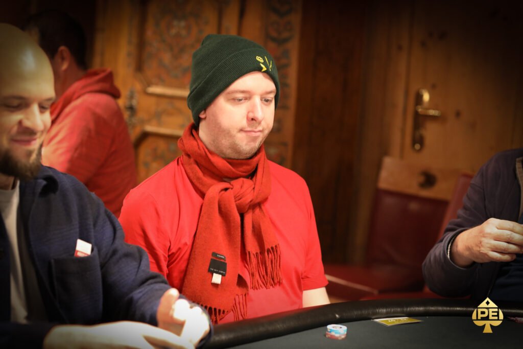 Ein Mann in einem schwarzen Anzug sitzt an einem Pokertisch und hält in seiner Hand eine Hand voll Spielkarten. Er hat einen konzentrierten Gesichtsausdruck und schaut auf die Karten. And