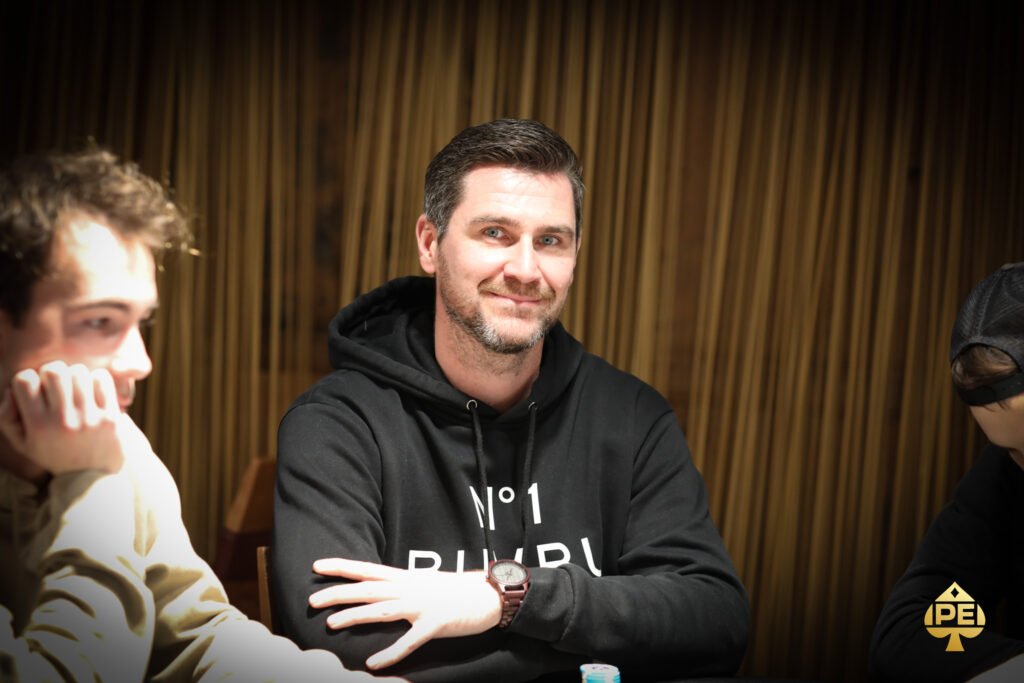 Ein Mann in einem blauen Anzug und einer Fliege sitzt an einem Pokertisch und hält seine Spielkarten in der Hand, während er konzentriert auf seine Gegner schaut. Im Hintergrund sind Poker