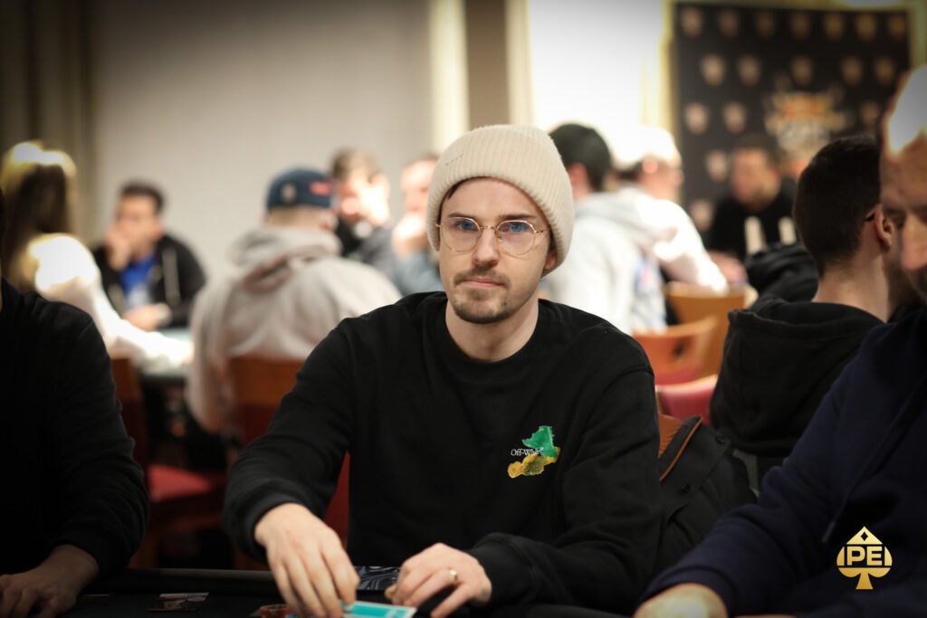 Ein Mann in einem schwarzen Anzug und mit einer Sonnenbrille sitzt an einem Pokertisch und betrachtet seine Karten konzentriert. Um ihn herum sind Pokerchips und Spielkarten verstreut.