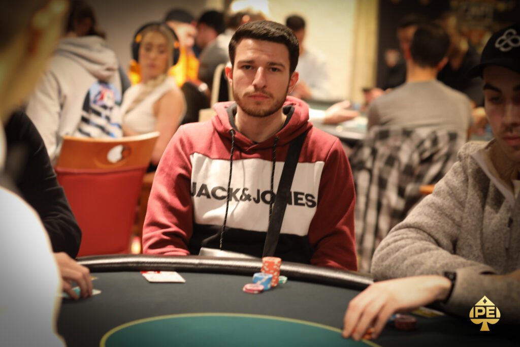 Ein Mann spielt Poker an einem Tisch mit grünem Filz. Er hat einen ernsten Gesichtsausdruck und hält Karten in der Hand. Im Hintergrund sind Pokerchips und Getränke zu