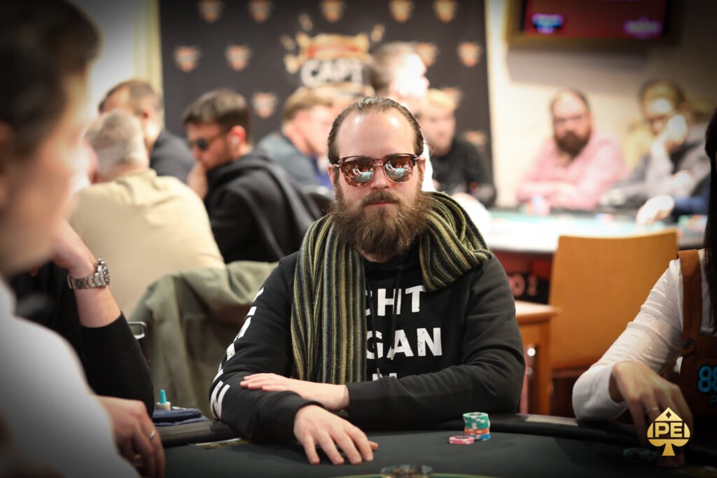 Ein Mann sitzt an einem Pokertisch und hält eine Hand mit Spielkarten. Er trägt einen dunklen Anzug und eine schwarze Krawatte. Um ihn herum sind Pokerchips und Getränke auf dem