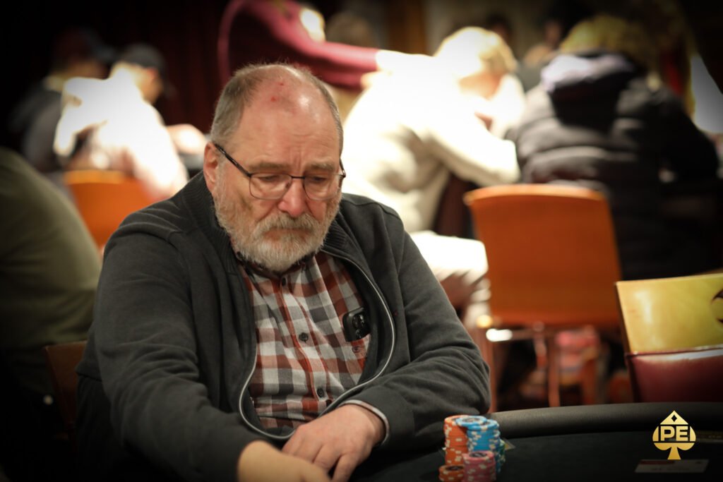 Ein Mann sitzt an einem Pokertisch und hält seine Karten mit ernstem Gesichtsausdruck. Im Hintergrund sind andere Spieler und Chips zu sehen.