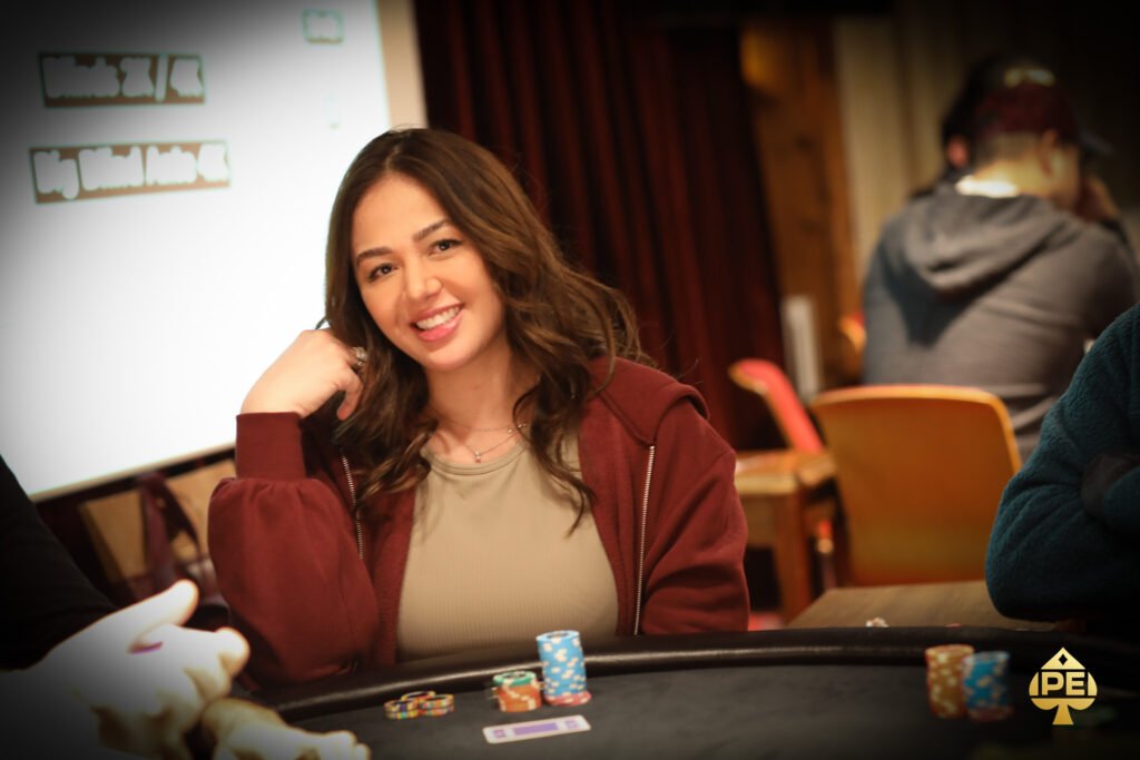 Ein Mann und eine Frau sitzen an einem Pokertisch und spielen Karten. Der Mann hat eine Kappe auf und schaut konzentriert auf seine Karten, während die Frau lächelt und ihre Karten hält. Im