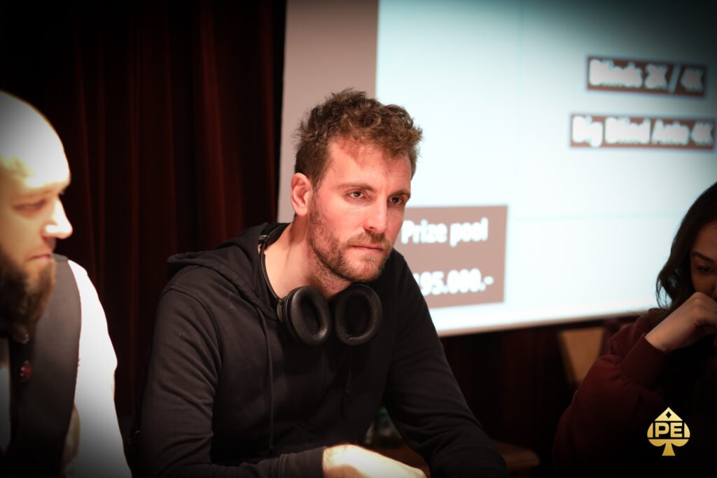 Ein Mann in einem schwarzen Anzug und einer weißen Hemdweste spielt Poker an einem eleganten Spieltisch, umgeben von anderen Spielern und einer glamourösen Atmosphäre.