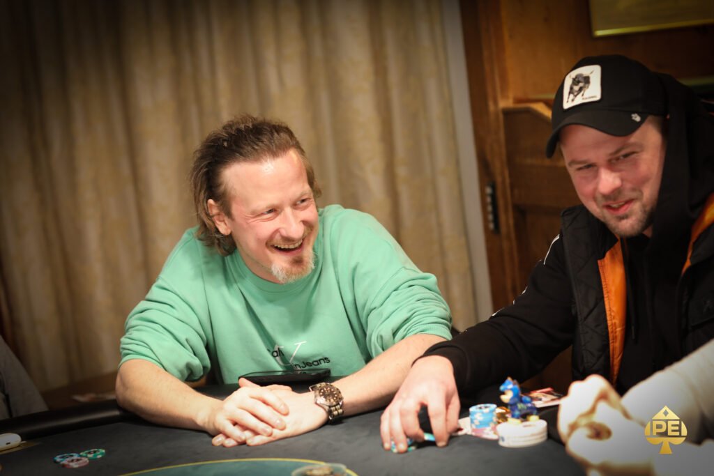 Ein Mann in einem dunkelblauen Anzug und einer Krawatte sitzt an einem Pokertisch und hält seine Karten in der Hand. Er wirkt konzentriert und fokussiert auf das Spielgeschehen