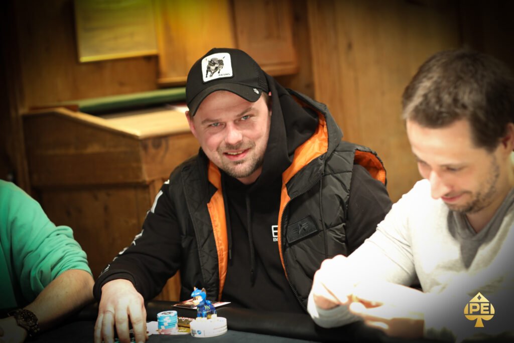 Ein Mann in einem dunkelblauen Anzug und einer hellblauen Krawatte sitzt an einem Pokertisch und betrachtet konzentriert seine Karten. Im Hintergrund sind Pokerchips und eine Glühlam