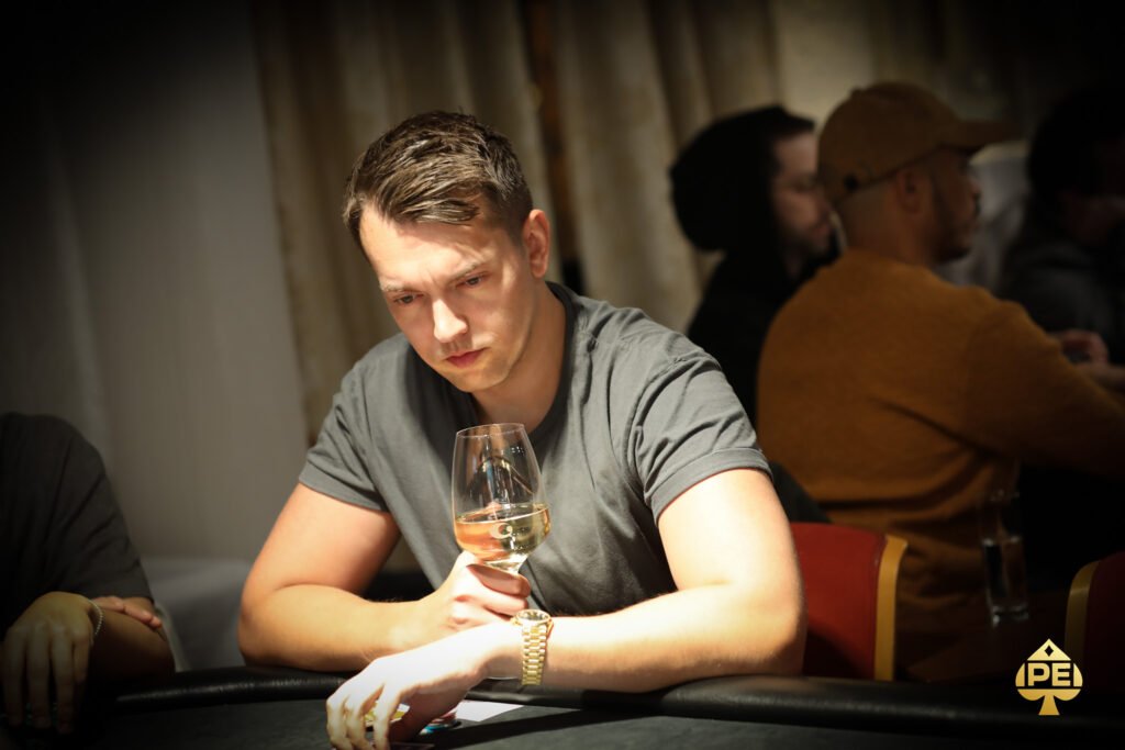Ein Mann in einem dunkelblauen Anzug sitzt an einem Pokertisch und hält seine Karten in der Hand, während er konzentriert auf seine Gegner schaut. Im Hintergrund sind Casino-Chips und Spiel