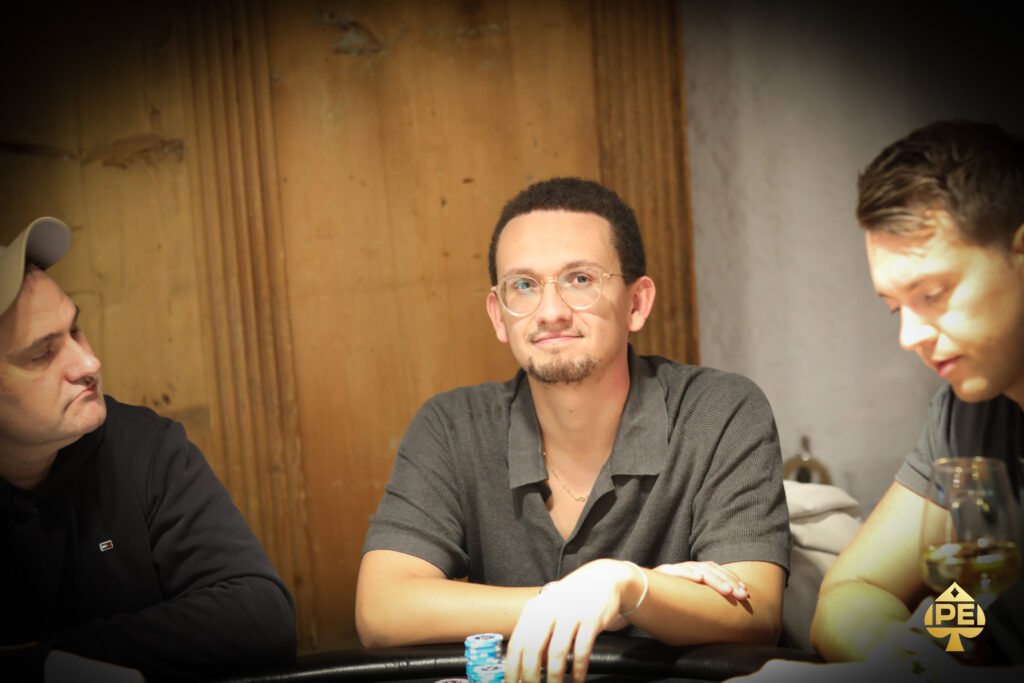 Ein Mann sitzt an einem Pokertisch und hält zwei Spielkarten in der Hand, während er konzentriert auf den Tisch schaut. Um ihn herum sitzen andere Spieler, die ebenfalls Karten halten. Die Atmos