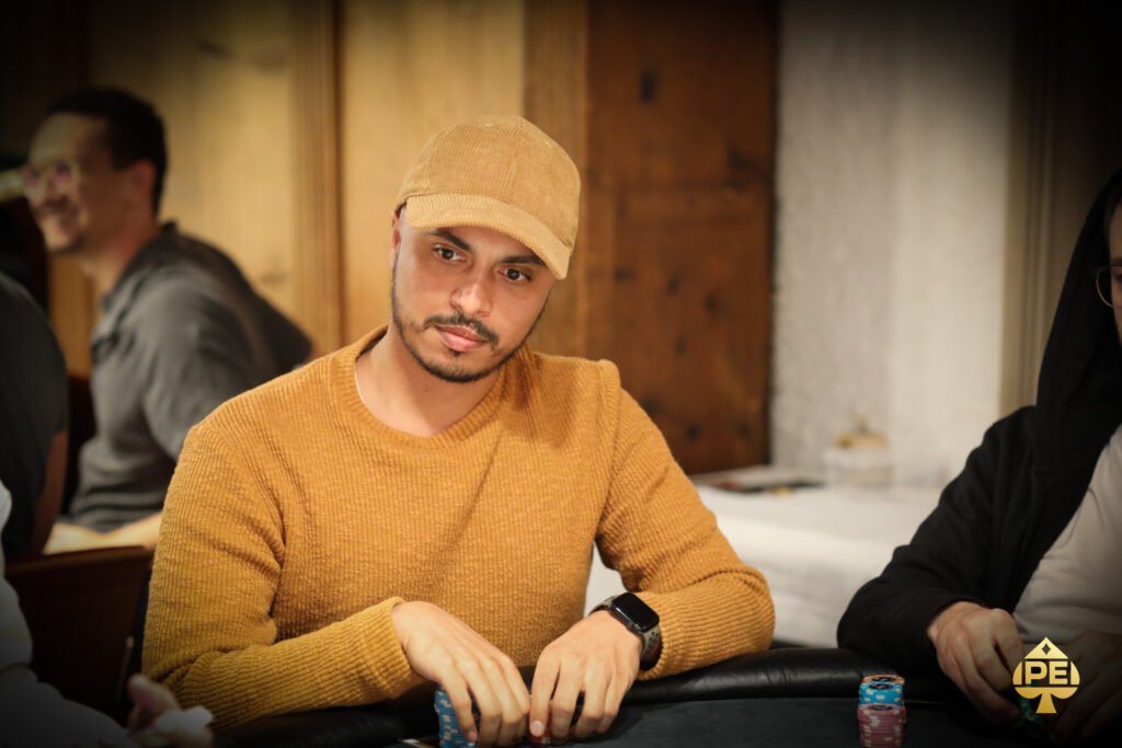 Ein Mann in einem dunkelblauen Anzug und einer weißen Hemd, der in einem eleganten Casino an einem Pokertisch sitzt und konzentriert auf seine Karten schaut. Im Hintergrund sind andere Spieler und