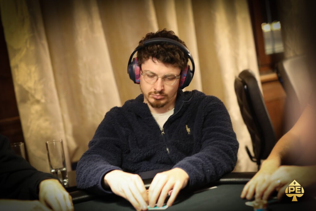Ein Mann sitzt an einem Pokertisch und hält zwei Spielkarten in der Hand. Er trägt einen Anzug und hat eine ernste Miene. Um ihn herum sind Pokerchips und Gläser zu sehen.