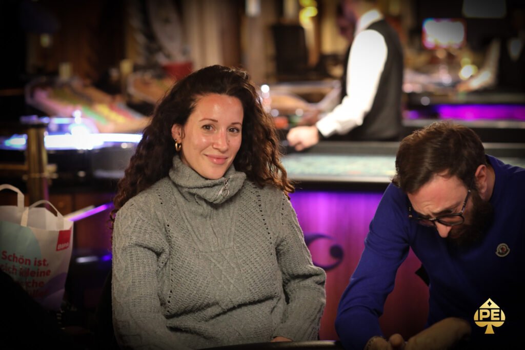 Ein Mann sitzt an einem Pokertisch und hält zwei Karten in der Hand, während er konzentriert auf seine Gegner schaut. Im Hintergrund sind Pokerchips und weitere Spieler zu sehen.