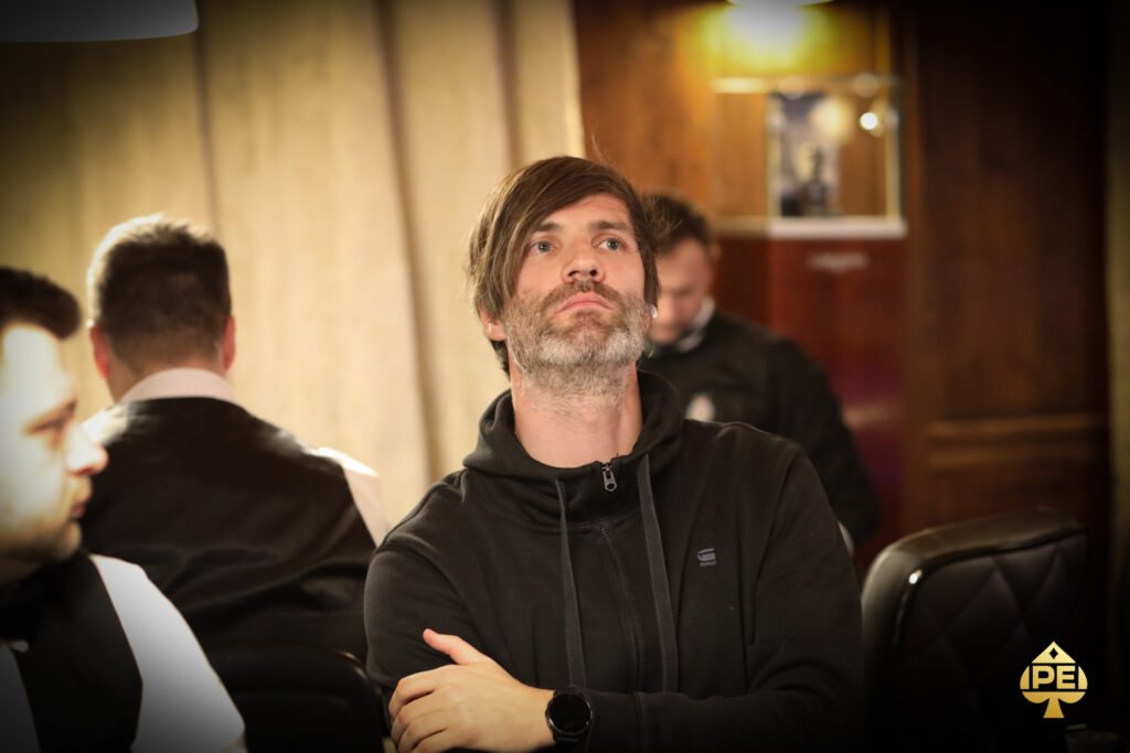 Ein Mann spielt Poker an einem Tisch mit grüner Filzdecke. Er hat eine Hand mit Spielkarten in der Hand und betrachtet sie konzentriert. Im Hintergrund sind Pokerchips und andere Spieler