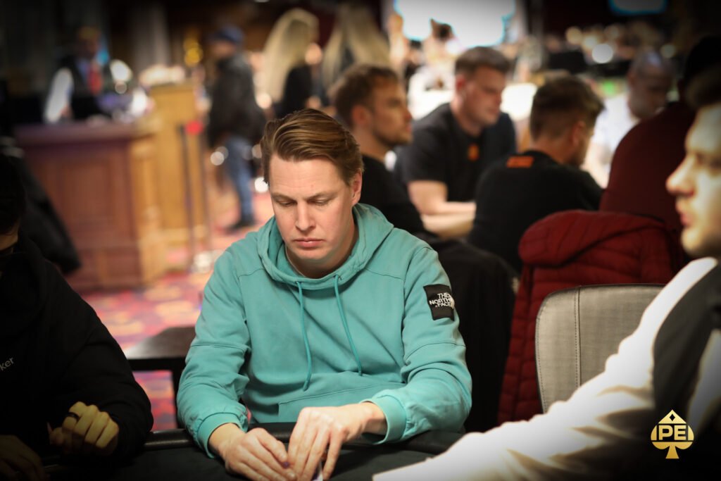 Ein Mann in einem schwarzen Anzug und einer schwarzen Sonnenbrille spielt Poker an einem Tisch mit grünem Filz. Er hält Karten in der Hand und hat einen ernsten Gesichtsausdruck.