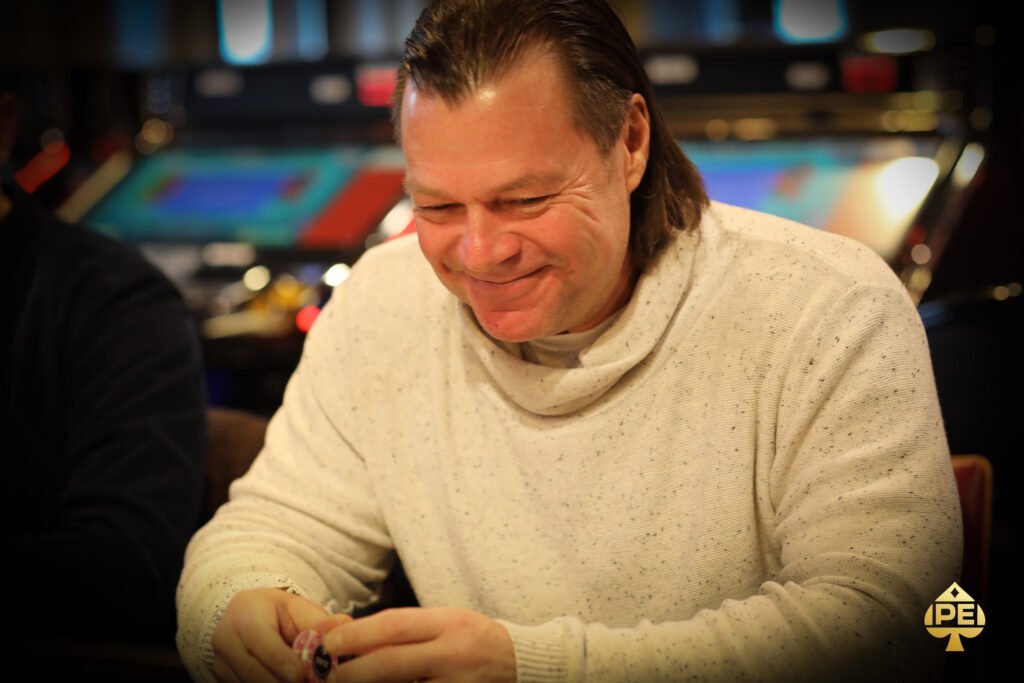 Ein Mann mit einer schwarzen Sonnenbrille und einem Hut sitzt an einem Pokertisch und hält seine Karten in der Hand, während er konzentriert auf den Tisch schaut. Andere Spieler sind im Hinter