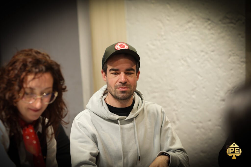 Ein Mann sitzt an einem Pokertisch und hält zwei Spielkarten in der Hand, während er konzentriert auf seine Gegner schaut. Die Szene ist in einem eleganten Pokerraum mit grünen Filz