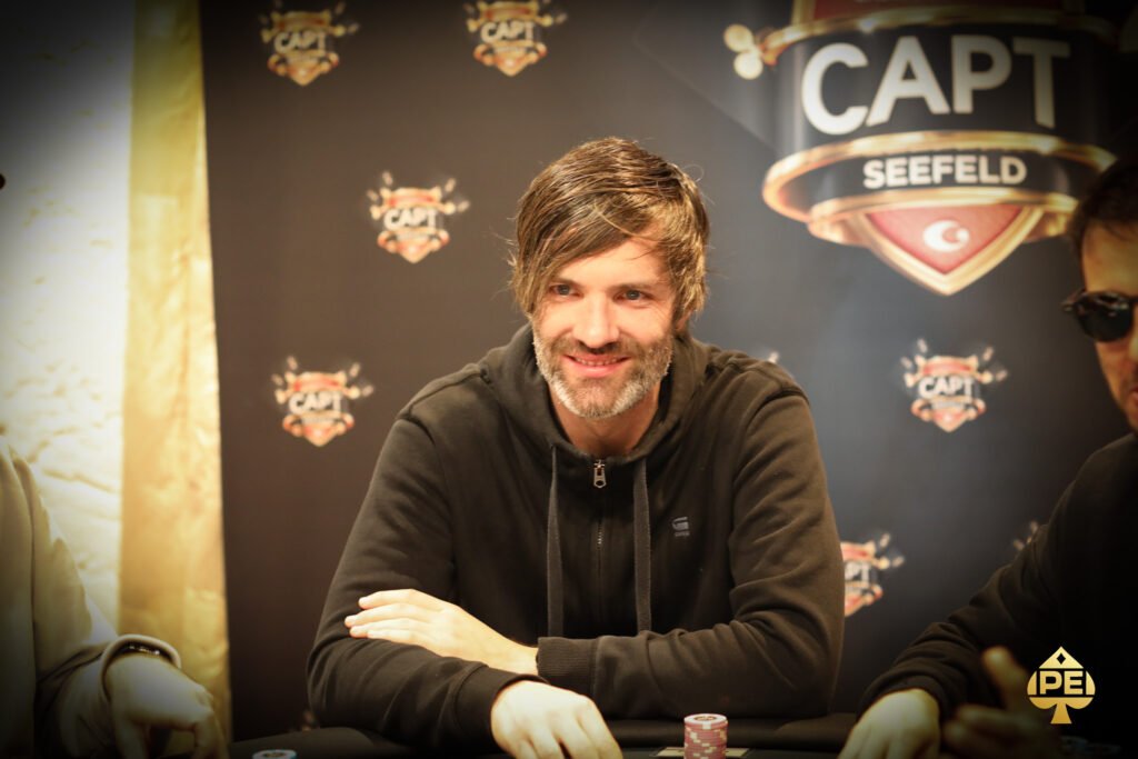 Ein Mann und eine Frau sitzen an einem Pokertisch und spielen Karten. Der Mann hat eine Hand am Kinn und schaut konzentriert auf seine Karten, während die Frau lächelnd ihre Karten betrachtet