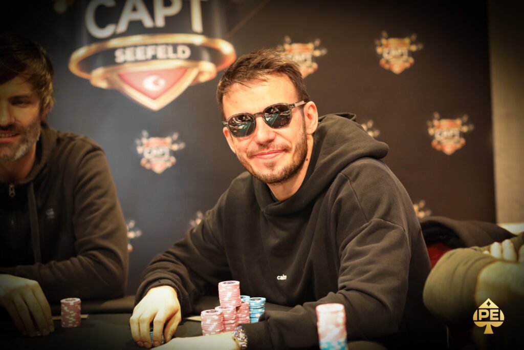 Ein Mann sitzt an einem Pokertisch und hält seine Karten verdeckt in der Hand. Er trägt eine dunkle Sonnenbrille und konzentriert sich auf das Spiel. Im Hintergrund sind andere Spieler und
