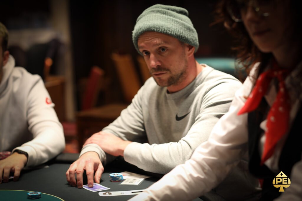 Ein Mann und eine Frau sitzen an einem Pokertisch und spielen Poker. Der Mann hat einen Stapel Chips vor sich liegen und hält eine Handkarte, während die Frau ihre Handkarten betrachtet. In der Mitte