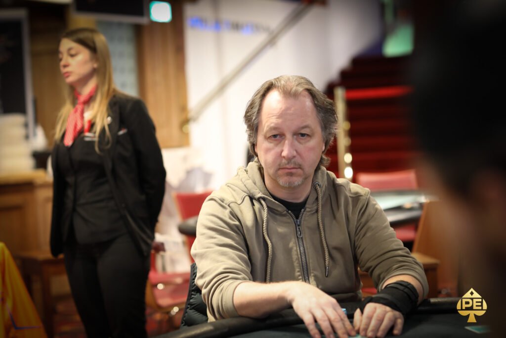 Ein Mann sitzt an einem Pokertisch und betrachtet konzentriert seine Karten. Im Hintergrund sind weitere Spieler und Chips zu sehen.