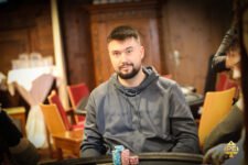Ein Mann in einem dunkelblauen Anzug und einer schwarzen Krawatte sitzt an einem Pokertisch und hält zwei Spielkarten in der Hand. Er wirkt konzentriert und schaut aufmerksam auf den