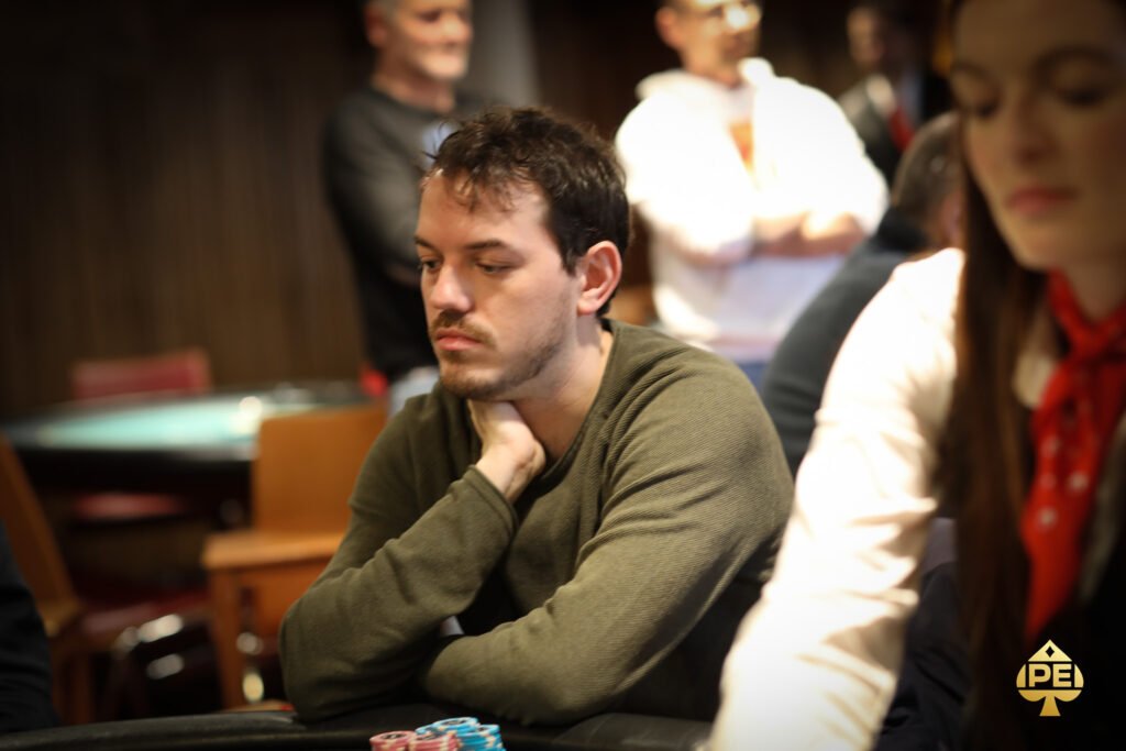 Ein Mann im Anzug sitzt an einem Pokertisch und betrachtet konzentriert seine Karten. Im Hintergrund sind andere Spieler und Pokerchips zu sehen.