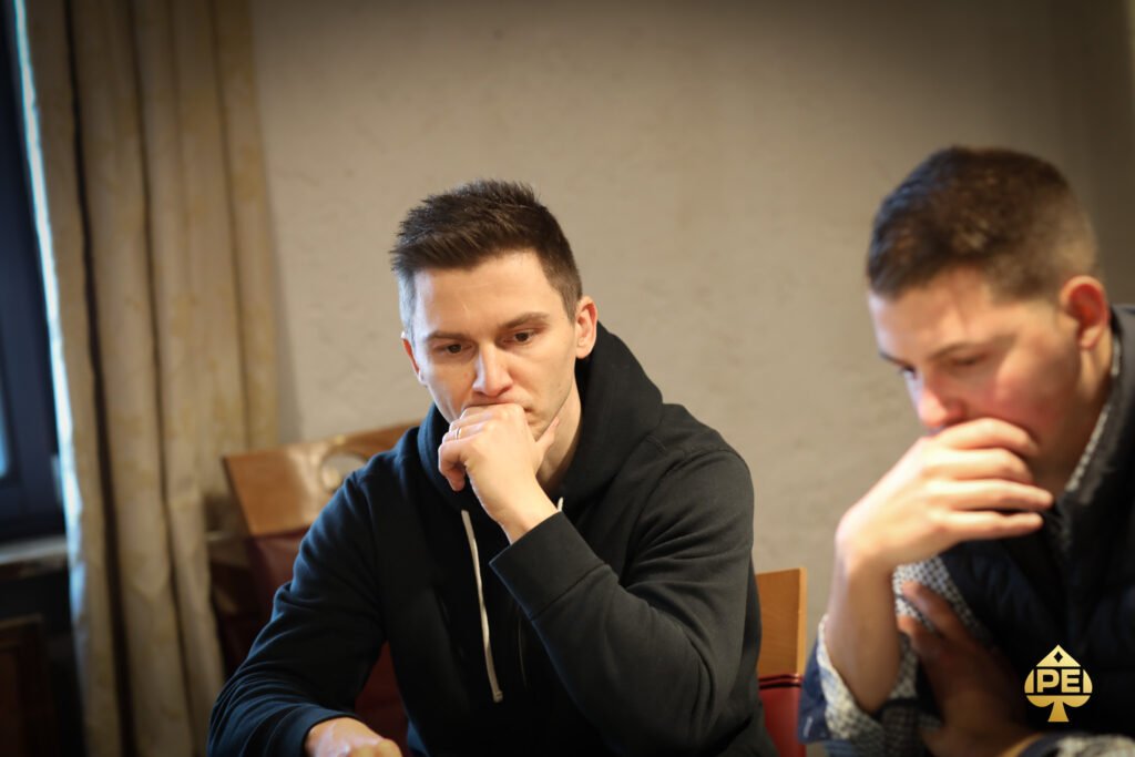 Ein Mann in einem schwarzen Anzug und einer blauen Krawatte steht vor einem Pokertisch mit Chips und Karten. Er hält eine Hand mit Karten und schaut konzentriert auf seine Gegner.