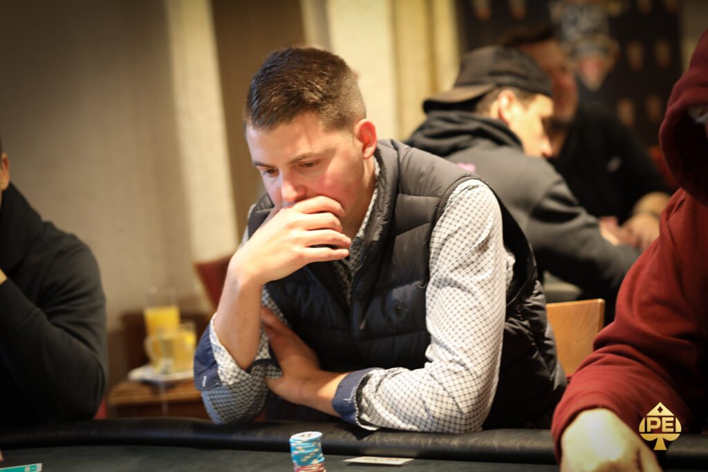 Ein Spieler hält zwei Spielkarten in der Hand und schaut konzentriert auf den Pokertisch, wo Pokerchips und weitere Karten liegen. Im Hintergrund sind andere Spieler und eine elegante Pokerrunde zu
