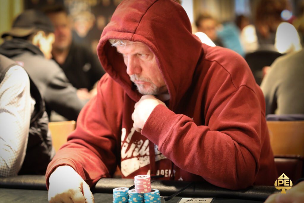 Ein Mann und eine Frau sitzen an einem Pokertisch, konzentriert auf ihre Karten und Chips. Im Hintergrund sind andere Pokerspieler und eine beleuchtete Bar zu sehen.
