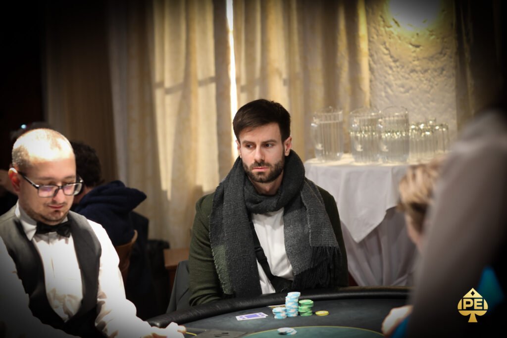 Ein Mann in einem dunkelblauen Anzug und einer weißen Hemdweste spielt Poker an einem eleganten Holztisch in einem luxuriösen Casino. Er hält seine Karten in der Hand und betrachtet sie