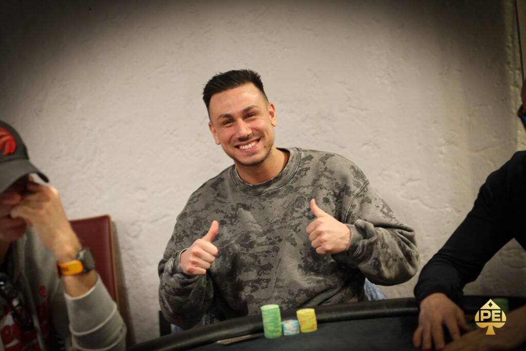Ein Mann sitzt an einem Pokertisch mit grünem Filz und hält zwei Spielkarten in der Hand. Er scheint konzentriert auf seine Hand zu schauen, während andere Spieler im Hintergrund verschwommen