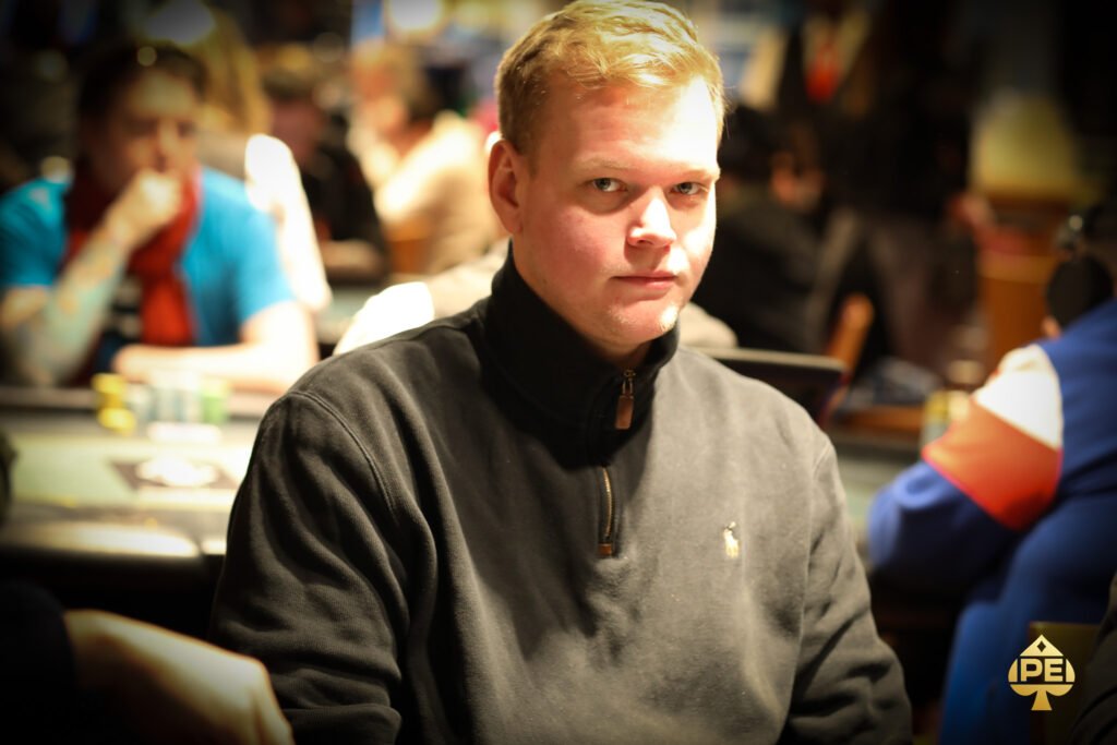 Ein Mann und eine Frau sitzen an einem Pokertisch und spielen eine Partie Poker. Der Mann hat einen Stapel Chips vor sich und hält eine Handkarte, während die Frau konzentriert ihre Karten betrachtet.