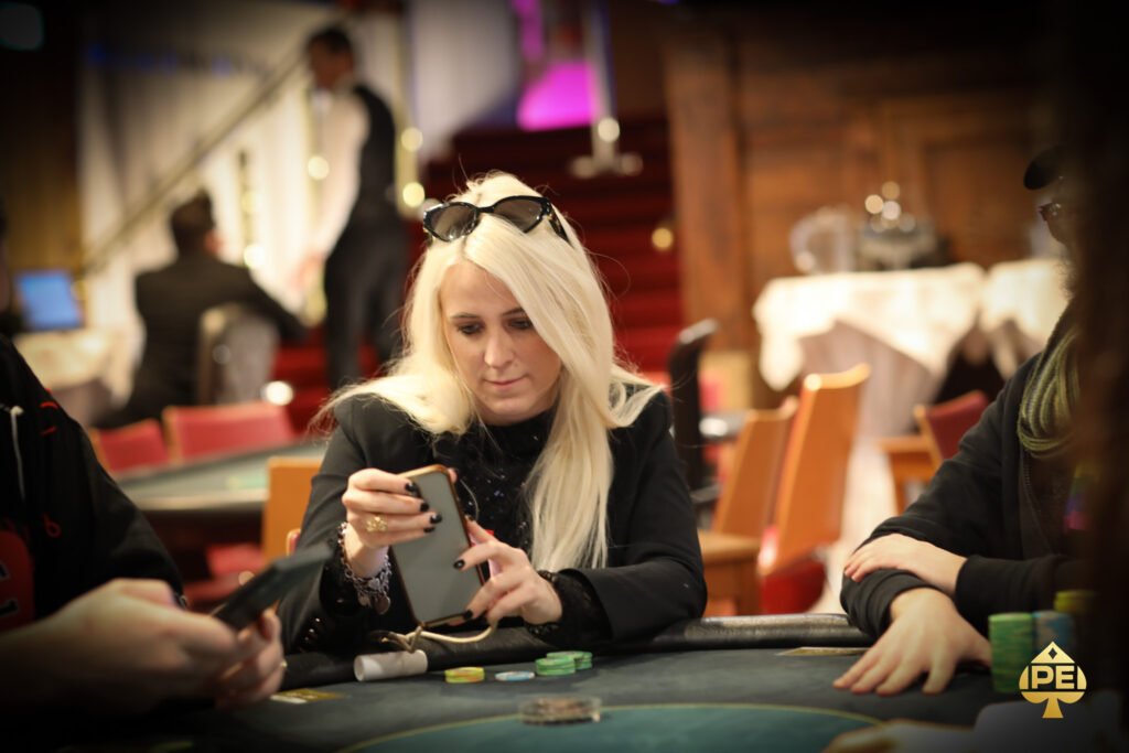 Ein Mann in einem eleganten Anzug sitzt an einem Pokertisch und hält zwei Spielkarten in der Hand. Er schaut konzentriert auf seine Karten, während im Hintergrund andere Spieler und Pokerchips zu