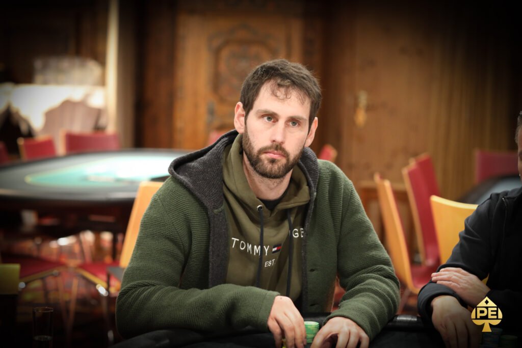 Ein Mann in einem schwarzen Anzug und einer schwarzen Sonnenbrille sitzt an einem Pokertisch und hält seine Spielkarten in der Hand. Im Hintergrund sind Casino-Chips und andere Spieler zu sehen.