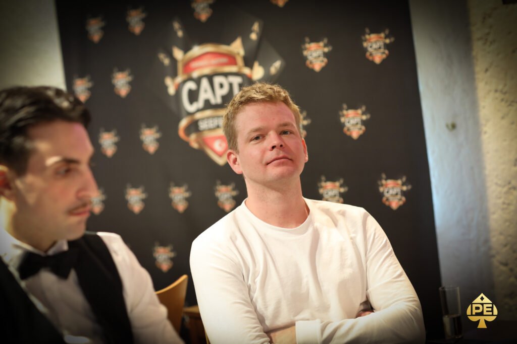 Ein Mann sitzt an einem Pokertisch und hält zwei Spielkarten in der Hand, während er konzentriert auf die Karten auf dem Tisch schaut. Um ihn herum sind Pokerchips und Gläser mit