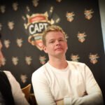 Ein Mann sitzt an einem Pokertisch und hält zwei Spielkarten in der Hand, während er konzentriert auf die Karten auf dem Tisch schaut. Um ihn herum sind Pokerchips und Gläser mit