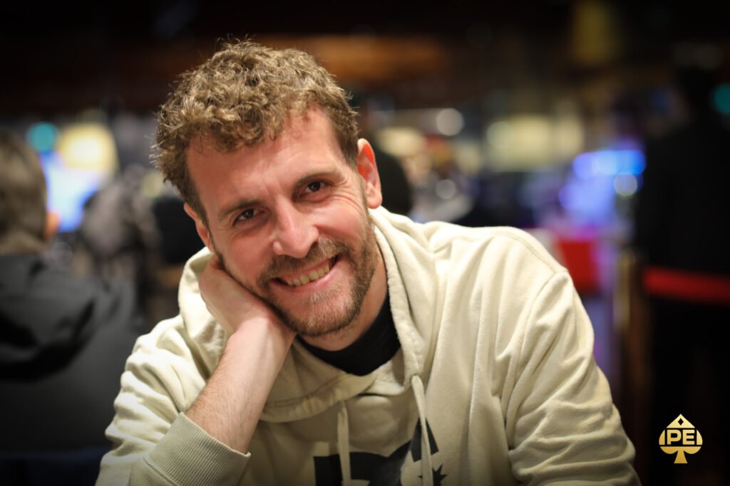 Ein Mann sitzt an einem Pokertisch und hält zwei Karten in der Hand. Er schaut konzentriert auf den Tisch, auf dem Pokerchips und Spielkarten liegen. Im Hintergrund sind weitere Spieler
