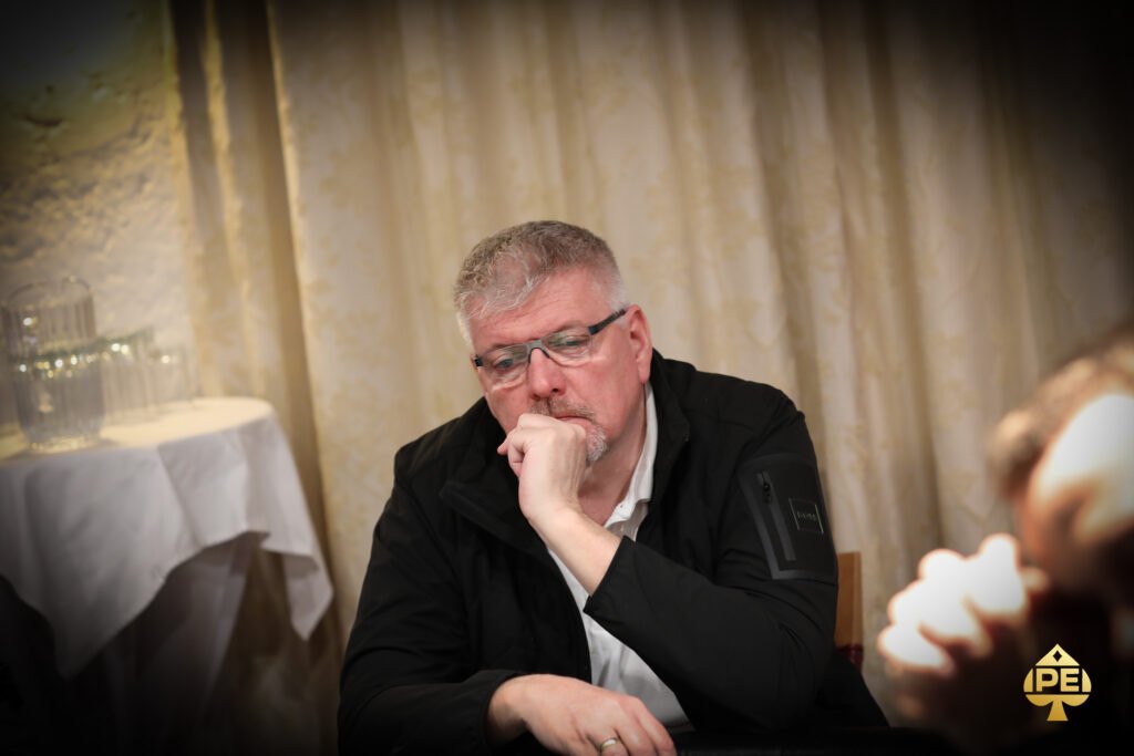 Ein Mann mit einem roten Hemd und einer schwarzen Weste sitzt an einem Pokertisch und hält seine Karten. Im Hintergrund sind weitere Spieler und Pokerchips zu sehen.
