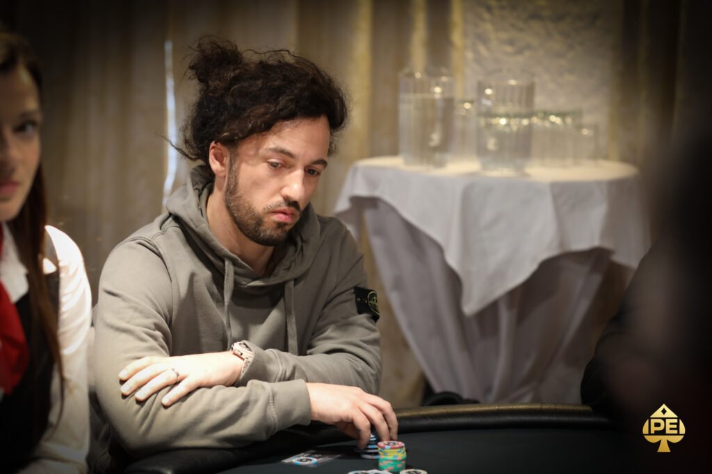 Ein Mann sitzt an einem Pokertisch und hält zwei Karten in der Hand. Er schaut konzentriert auf den Tisch, auf dem Pokerchips und Spielkarten liegen. Im Hintergrund sind weitere Spieler