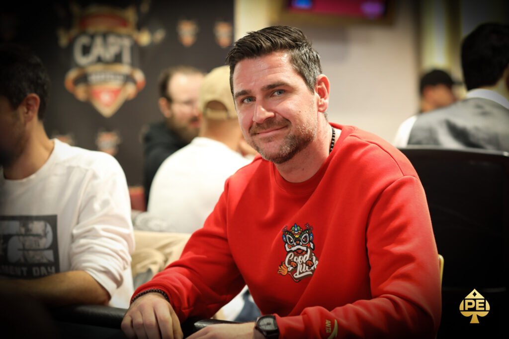 Ein Mann spielt Poker an einem Tisch mit grüner Filzdecke, umgeben von anderen Spielern. Er hält Karten in der Hand und hat einen konzentrierten Ausdruck auf seinem Gesicht.