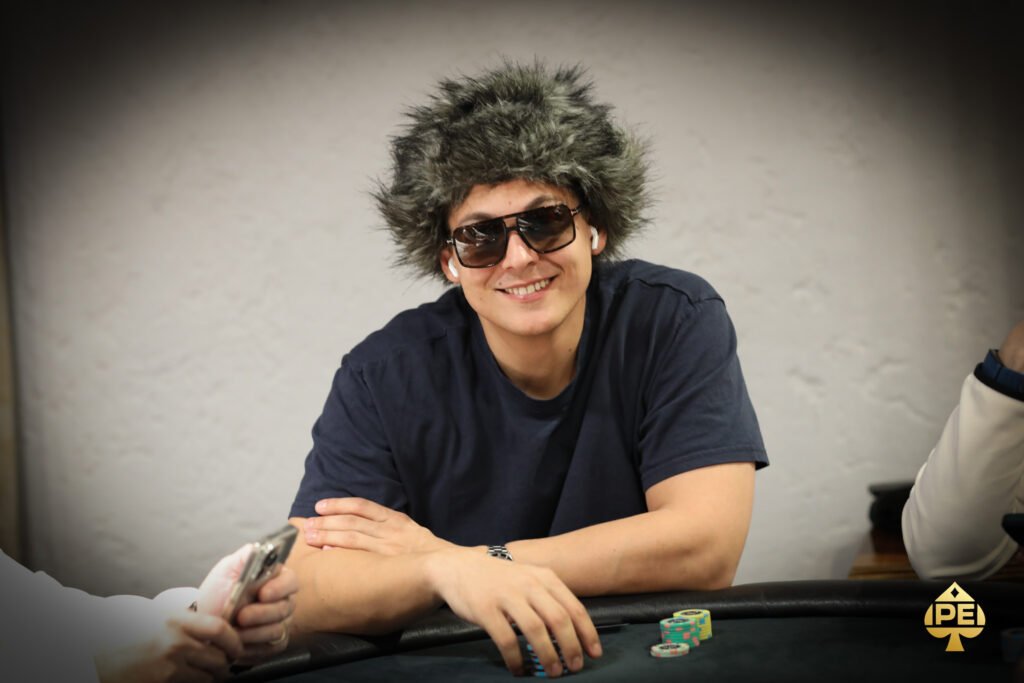 Ein Mann in einem schwarzen Anzug und einer schwarzen Sonnenbrille sitzt an einem Pokertisch und hält zwei Karten in der Hand. Er schaut konzentriert auf die Karten und wirkt entschlossen
